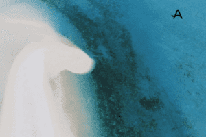 uftaufnahme eines weißen Sandstrandes mit sanft geschwungener Küstenlinie, die ins türkisfarbene Meer übergeht. Dunklere Bereiche im Wasser deuten auf Korallen oder Seegras hin. In der oberen rechten Ecke befindet sich ein stilisiertes "A".
