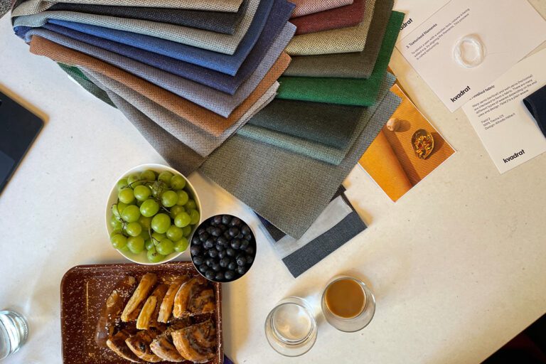 Nahaufnahme verschiedener Stoffmuster von Kvadrat in abgestuften Farben – von Blau über Beige bis Weinrot – auf einem Tisch mit einem Wasserglas, Stift und einer grob gewebten Stoffprobe.