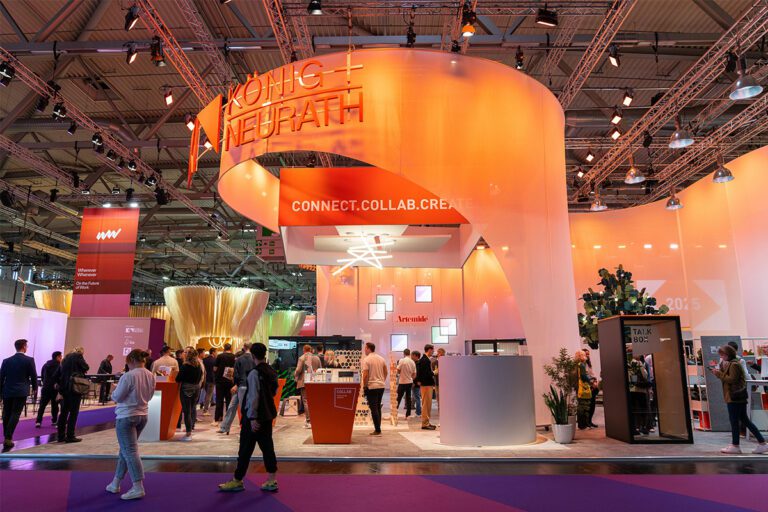 Das Bild zeigt den eindrucksvollen Messestand von König + Neurath anlässlich des 100-jährigen Jubiläums des Unternehmens. Unter dem Motto „CONNECT. COLLAB. CREATE.“ werden innovative Lösungen für die Zukunft der Arbeit präsentiert. Der Stand ist modern, offen und einladend gestaltet – mit einer gelungenen Mischung aus Architektur, Licht und Markeninszenierung. Ein starkes Statement für Designkompetenz und Teamarbeit.