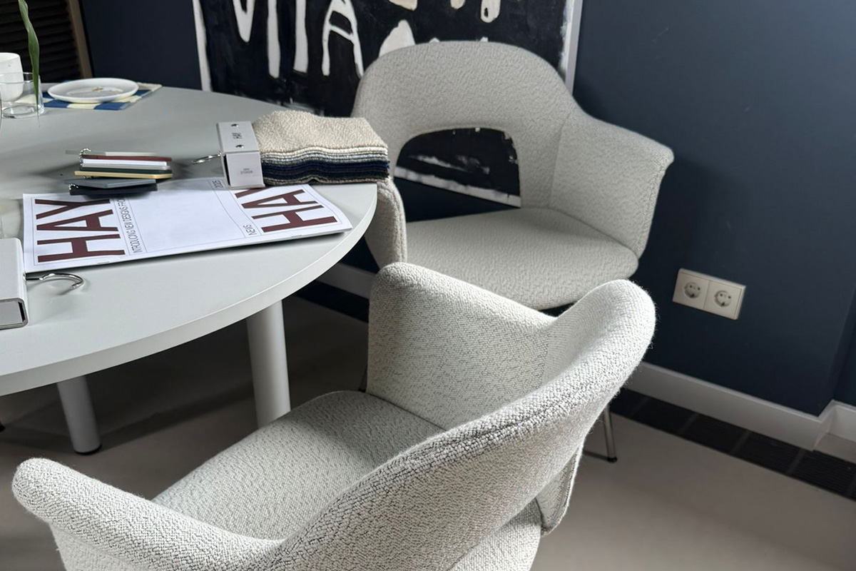 Zwei moderne, weiß strukturierte Sessel stehen um einen hellgrauen, runden Tisch, auf dem HAY-Kataloge, Stoffmuster, ein Teller und Musterordner liegen. Die Szene wirkt wie ein Design-Showroom oder ein kreativer Arbeitsbereich mit dunkelblauer Wand, abstrakter Schwarz-Weiß-Kunst und sichtbaren Steckdosen im Hintergrund.