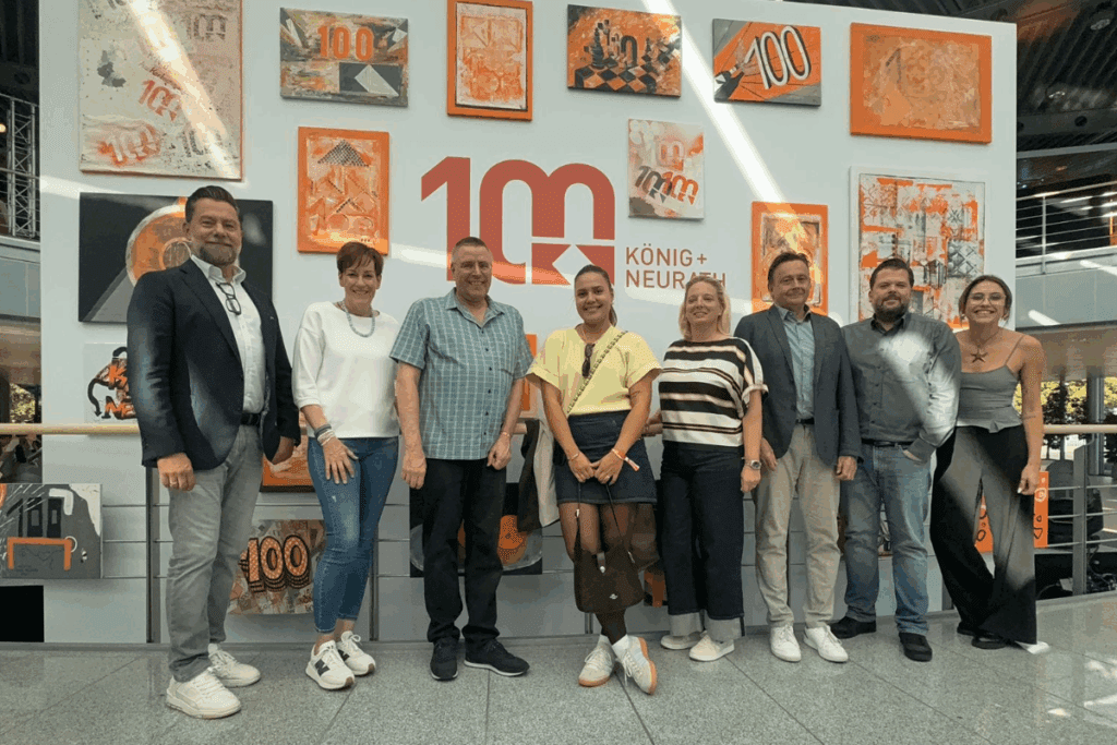 Neun Personen stehen lächelnd vor einer Wand mit gemalten Kunstwerken und dem zentralen Logo „100 Jahre König + Neurath“, das in Rot und Orange gestaltet ist.