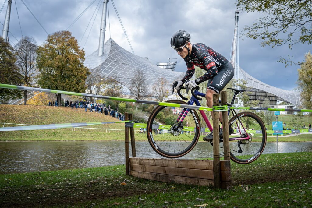 Radfahrer im Syntace-Teamtrikot springt mit dem Fahrrad über ein Hindernis im Olympiapark München, Zuschauer und Olympiadach im Hintergrund.