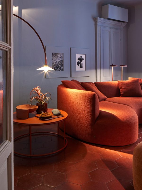 Ein elegantes, geschwungenes Cor Sofa in warmem Ziegelrot steht auf einem rotbraunen Fliesenboden, beleuchtet von einer stilvollen Bogenlampe. Daneben ein runder Beistelltisch mit minimalistischer Deko, eingebettet in eine stimmungsvolle Wohnzimmerszene mit moderner Kunst an der Wand.