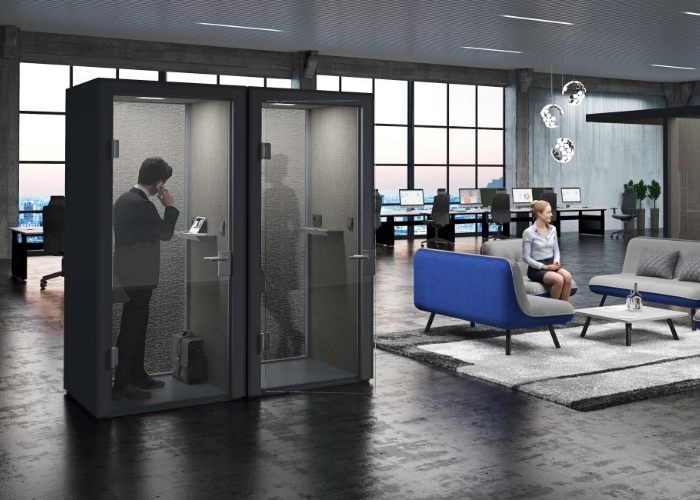 Das Bild zeigt eine moderne Büroumgebung, wo zentral zwei Telefonzellen von König+Neurath stehen. Die Telefonzellen haben Türen aus Glass. Im Hintergrund ist ein kleiner Loung Bereich in den Farben blau, grau gehalten, und eine Schreibtischreihe vor einen riesigen Fenster.