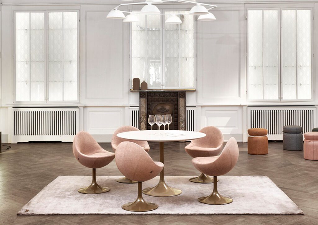 Ein großer Raum mit heller Wand und großen Fenstern. Mittig ein runder weißer Tisch und rum herum fünf Sessel in runder Form in Beigerosa und braunen Gestell.