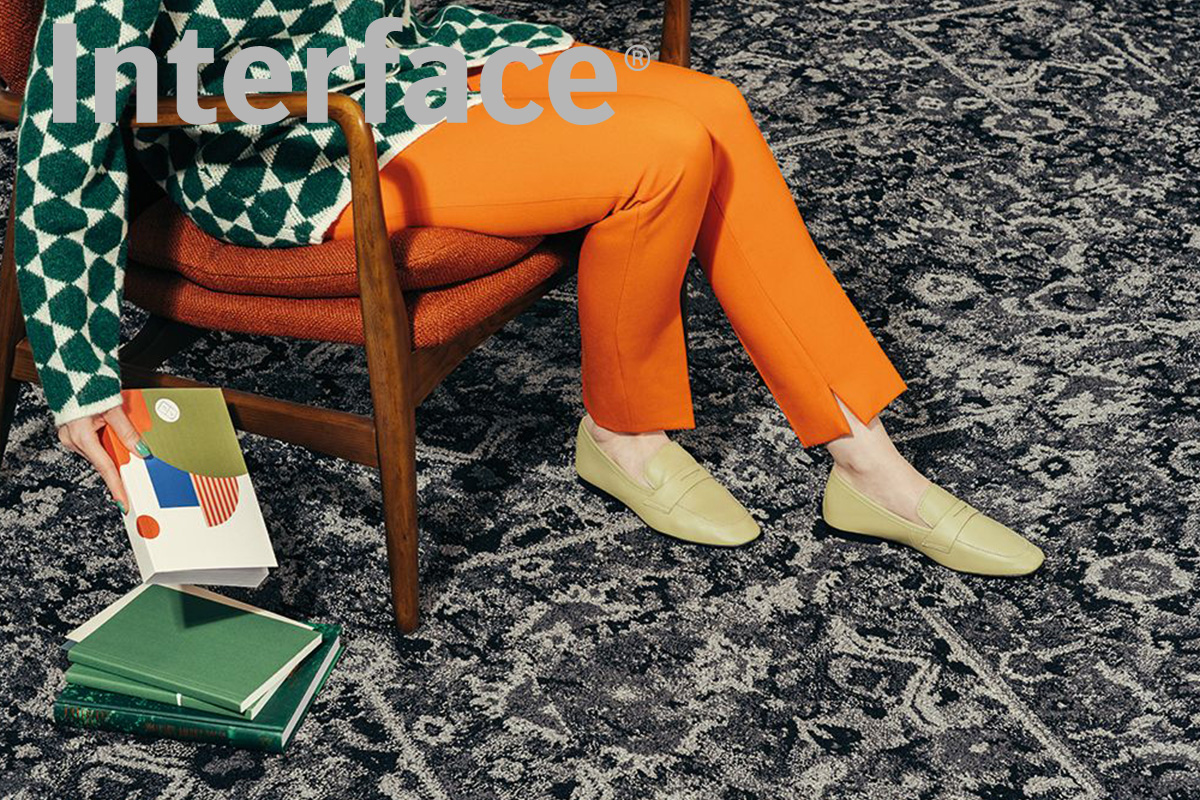 Eine Frau sitzt auf einem Holzhocker und trägt eine orangefarbene Hose. Sie hält ein Buch in der Hand und befindet sich auf einem Teppich mit verschiedenen Nostalgiemustern in Grautönen aus der 'Past Forward'-Kollektion von Interface.