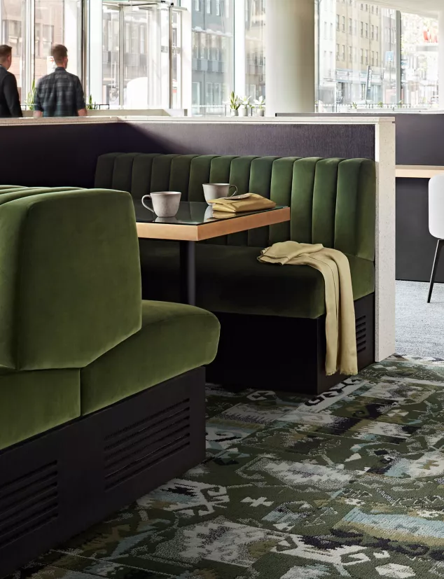 Der Raum ist mit einem Teppich der 'Past Forward' Kollektion von Interface gestaltet, der grüne, hellgraue und weiße Elemente vereint. Eine grüne Couch und ein Tisch bilden den zentralen Bereich, während sich eine weitere große Couch parallel dazu erstreckt.