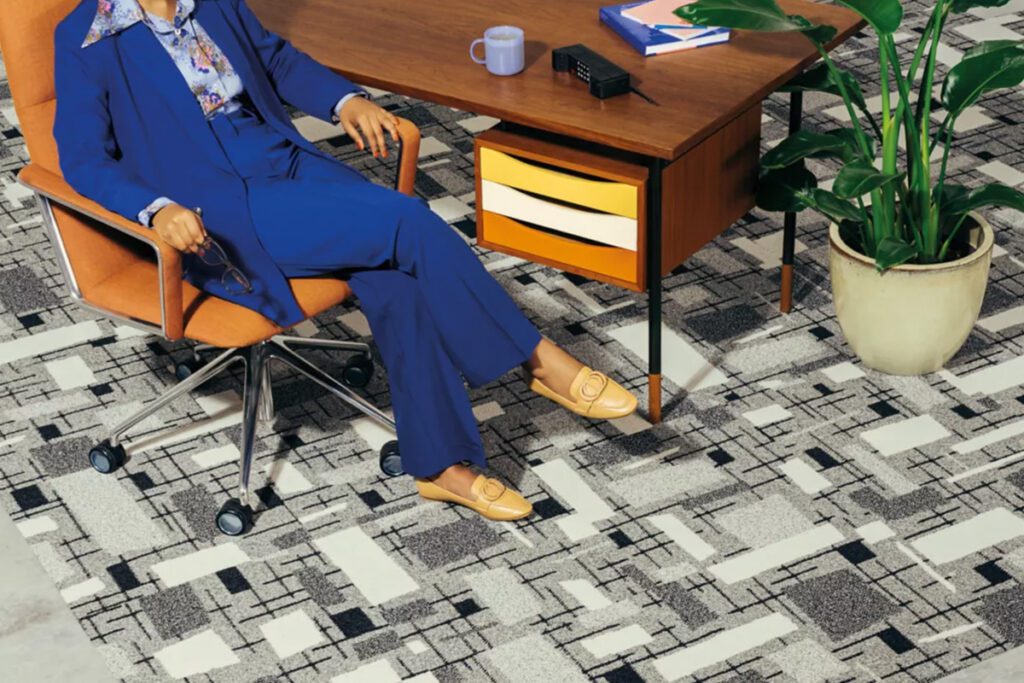 Ein Teppich von Interface in hellgrau, mit Vierecken in verschiedenen Grautönen, erstreckt sich über den Raum. Eine Frau sitzt in einem Rollstuhl an einem Schreibtisch, sie trägt ein blaues Sakko