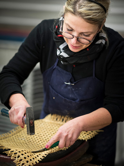 Eine Frau mit blonden Haaren und Brille befestigt das Rattangeflecht auf einem Holzgestell.