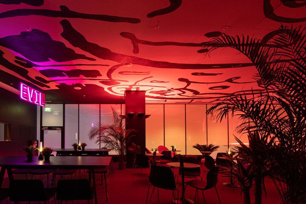 Inspirierender Live Jazz Club "Live Evil" in München. Rotes Licht taucht den Raum, hohe Wänden, eine detailreich gestaltete Decke. In der Mitte erstreckt sich eine Bar, während auf einer erhöhten Plattform mit Teppich Lounge-Möbel für entspannte Atmosphäre sorgen