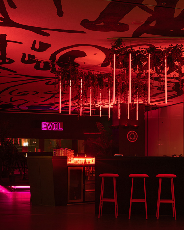 Andere Perspektive der beeindruckenden Bar im Jazzclub: Die leuchtende Bar in der Mitte des Raums ist von hohen, roten Hockern umgeben. Dahinter erstreckt sich das Podest mit der Leuchtreklame "Evil". Von diesem Blickwinkel aus fangen leuchtende Hängelampen an der Decke die Aufmerksamkeit ein, während Pflanzen stilvoll zwischen den Elementen platziert sind. Eine faszinierende Aussicht auf Stil und Atmosphäre im Live Jazz Club