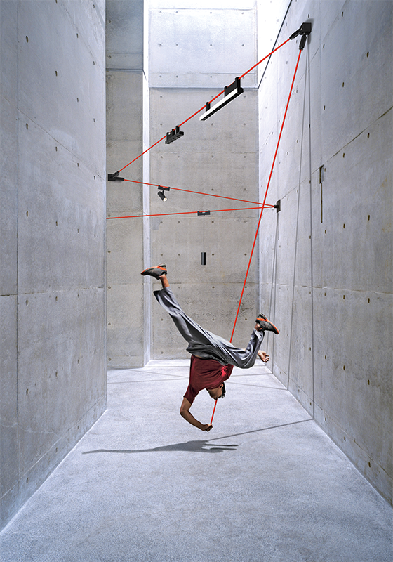 Ein Mann klettert an einem hellgrauen Raum entlang, in dem das Funivia Leuchtensystem von Artemide in Rot aufgespannt ist, ähnlich einer Seilbahn.