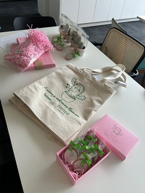 Ein Büroraum mit einem Tisch, auf dem Werbeartikel für eine Veranstaltung zu Valentinstag zu sehen ist, darunter eine Baumwolltasche mit dem Aufdruck 'Es müssen ja nicht immer Rosen sein', rosa Verpackungsmaterial, und eine offene Geschenkbox mit Keksen, alle gebrandet mit einem Kaktus und den Logos von Hauser und Zilenzio