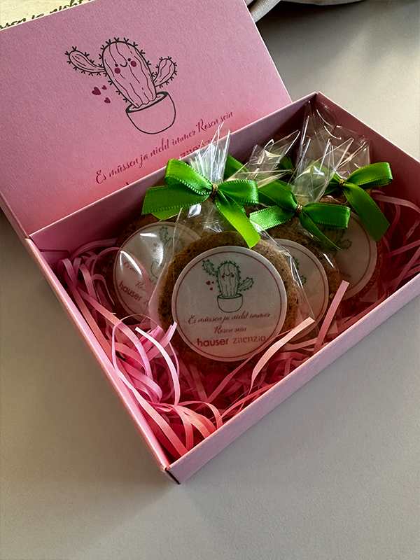 Eine offene, rosa Geschenkbox mit sechs einzeln verpackten Keksen, gebunden mit grünen Schleifen, auf rosa Sizzle-Pak. Die Innenseite des Deckels zeigt eine Kaktus-Illustration und den Text 'Es müssen ja nicht immer Rosen sein' sowie die Logos von Hauser und Zilenzio.