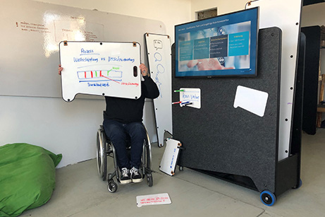 erson im Rollstuhl präsentiert auf einem mobilen Imsinne Whiteboard einen Prozessablauf, umgeben von weiteren flexiblen Whiteboard-Lösungen in einem hellen Workshop-Raum, was die barrierefreie Zugänglichkeit der Imsinne Produkte unterstreicht.