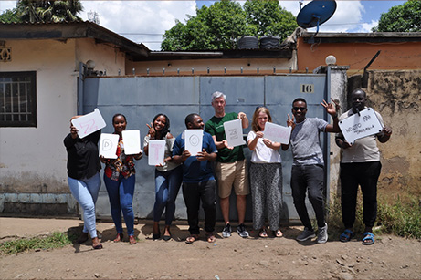 Eine Gruppe von Menschen, darunter Tobias, der Besitzer von Imsinne, steht vor einem Gebäude in Tansania und präsentiert Imsinne Whiteboards mit Buchstaben, die eine Botschaft bilden, und demonstriert die Gemeinschaft und Zusammenarbeit, die durch Imsinne Produkte ermöglicht werden.