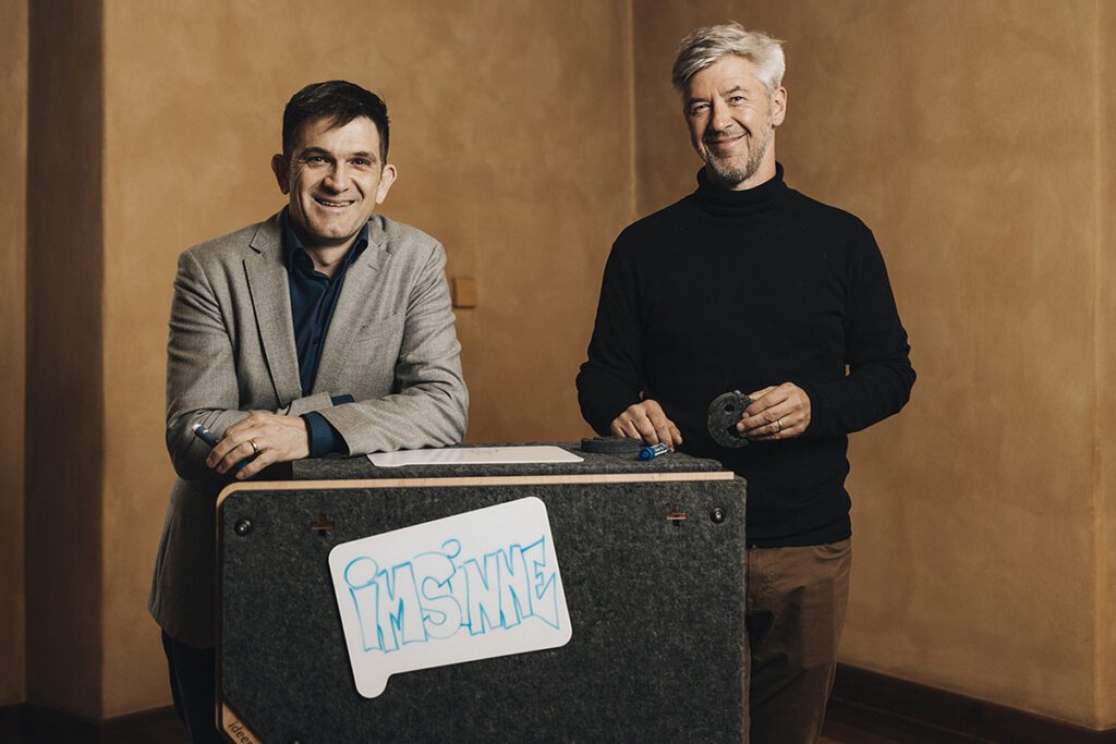 Tobias und Jonathan, die Gründer von Imsinne, stehen lächelnd neben einem mobilen Whiteboard des Unternehmens, das mit dem Imsinne-Logo gekennzeichnet ist, was die persönliche Verbindung und das Engagement hinter der Marke Imsinne hervorhebt.