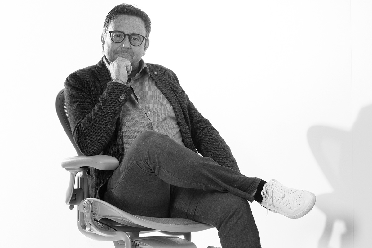 Reinhard Pelzer von Hauser mit Hemd, Sakko und Brille schaut in die Kamera und stützt sein Kinn mit einer Hand. Vor einem Weißen Hintergrund auf einem Bürostuhl sitzend, der im Hintergrund einen Schatten wirft.