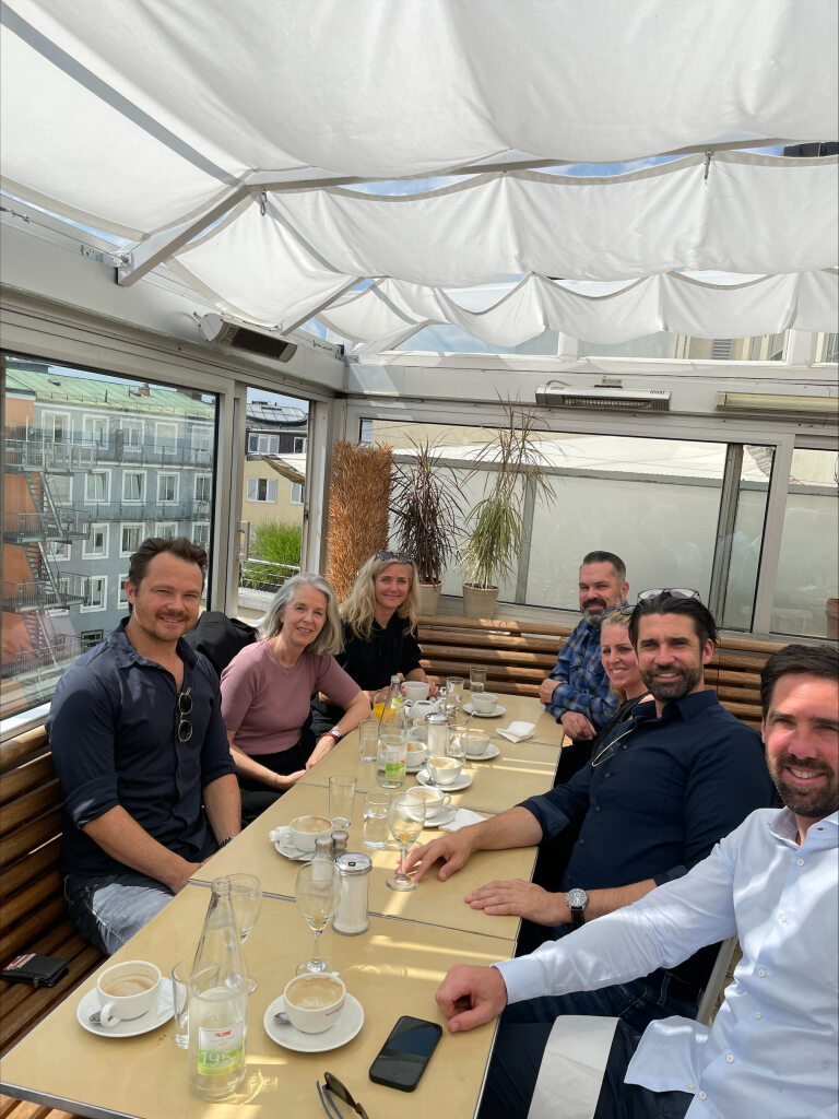 Eine Gruppe von sieben Personen sitzt um einen Tisch und genießt ein Frühstück in einem hellen Raum mit großen Fenstern und Pflanzen im Hintergrund. Alle lächeln und blicken in die Kamera, was eine entspannte und freundliche Atmosphäre vermittelt.