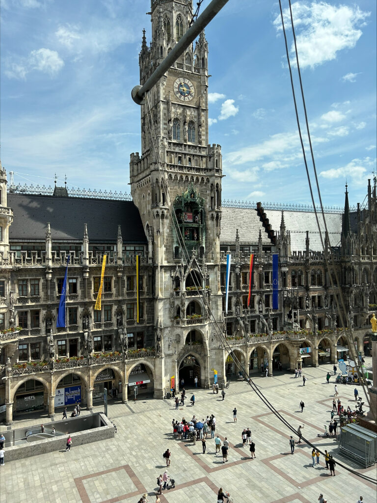 Blick auf den Marienplatz in München mit dem Neuen Rathaus im Hintergrund. Der Platz ist von Menschen belebt, und die Frauenkirche mit bunten Fahnen geschmückt. Der Himmel ist blau mit einigen Wolken.