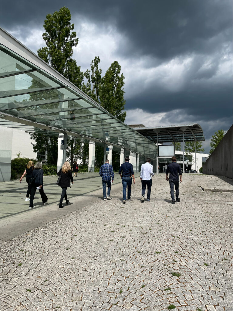 Mehrere Personen gehen unter einem Glasdach auf dem Außengelände der Messe Architect@Work in München. Der Himmel ist bewölkt, und im Hintergrund sind hohe Bäume zu sehen.