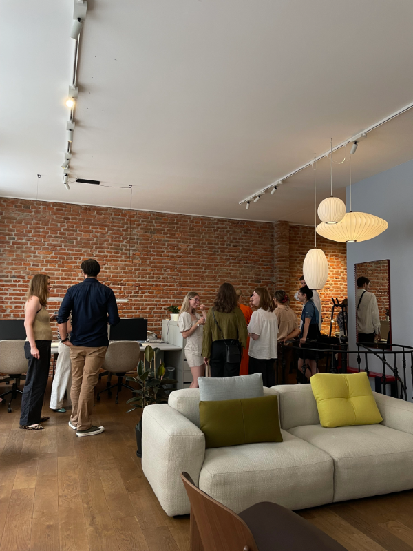 Ein weiterer Blick auf den Showroom mit Menschen, die sich in der Nähe eines gemütlichen Sitzbereichs und einer Ziegelakzentwand unterhalten. Moderne Beleuchtung und Möbel sind prominent zu sehen. Event im Hay Showroom München mit Hauser Office Design und Architekten.