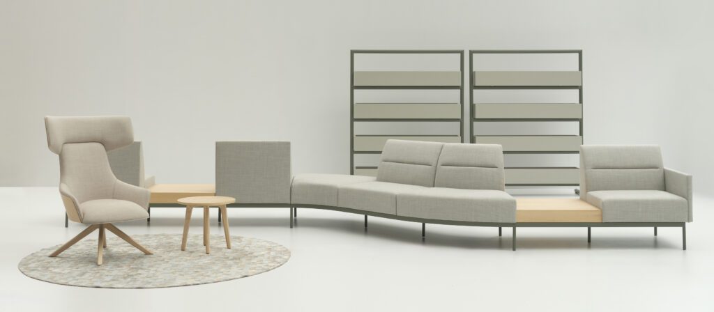 Hendix Lounge-Setup mit modularem Sofa, hellem Sessel und minimalistischen Beistelltischen auf einem geometrischen Teppich, ergänzt durch moderne Regale im Hintergrund.