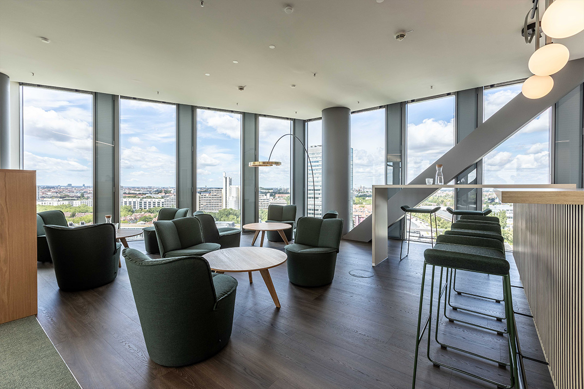 Ein Loungebereich mit grünen Sesseln und runden Holztischen, der mit einer Bar und hohen Stühlen kombiniert ist. Die großen Fenster bieten eine weitläufige Aussicht.