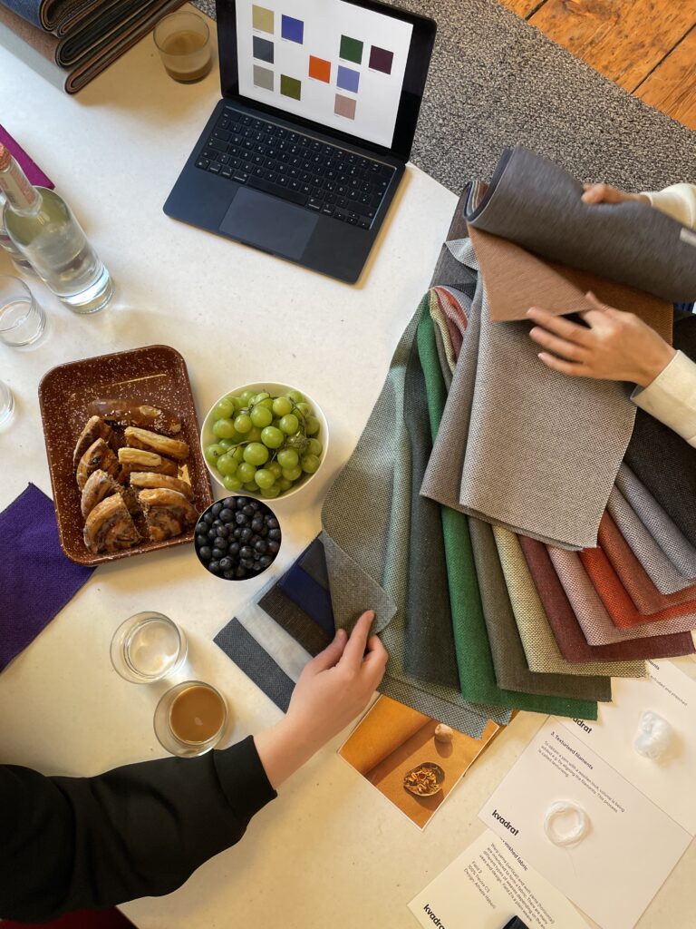 Zwei Personen betrachten Stoffmuster an einem Tisch, umgeben von Snacks, Getränken und einem Laptop mit Farbpalette – eine kreative Auswahl-Szene mit grünem, blauem und braunem Textil.