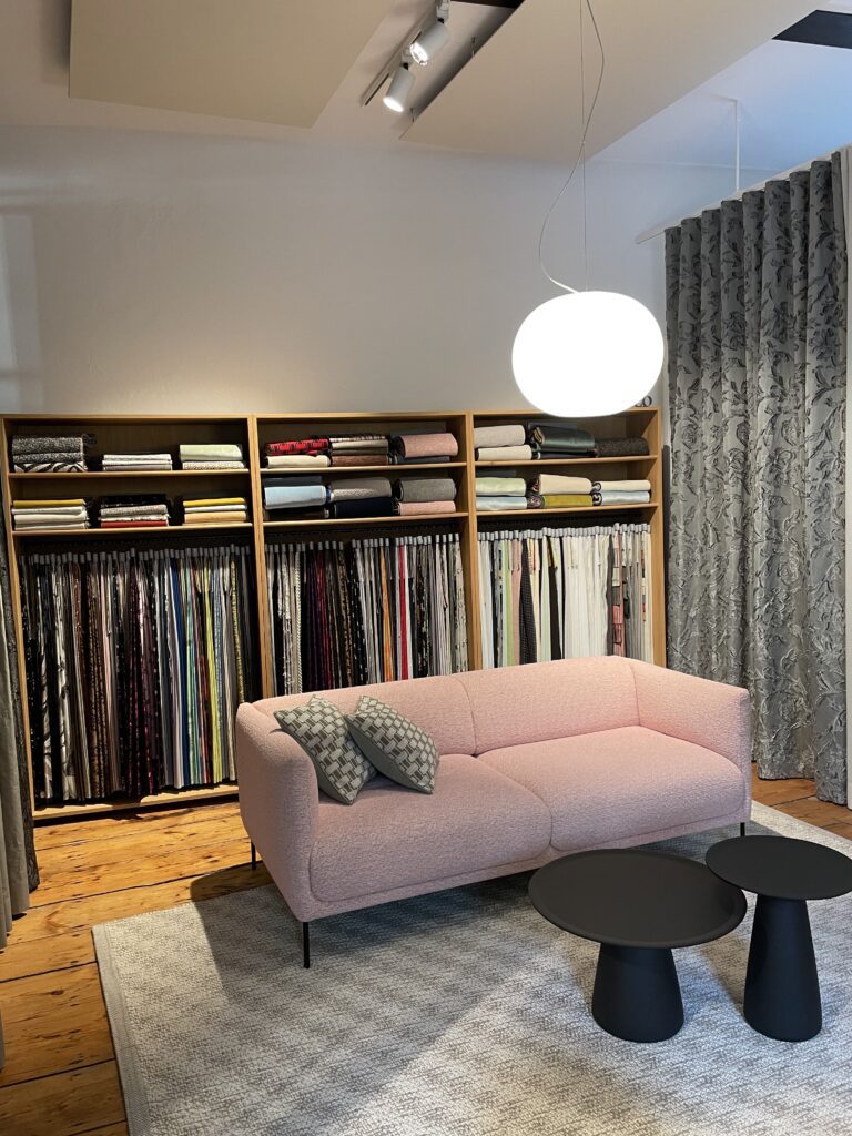 Ein stilvoll eingerichteter Showroom mit einem rosa Sofa, zwei gemusterten Kissen und schwarzen Beistelltischen, dahinter ein Regal voller Stoffrollen und hängender Stoffproben.