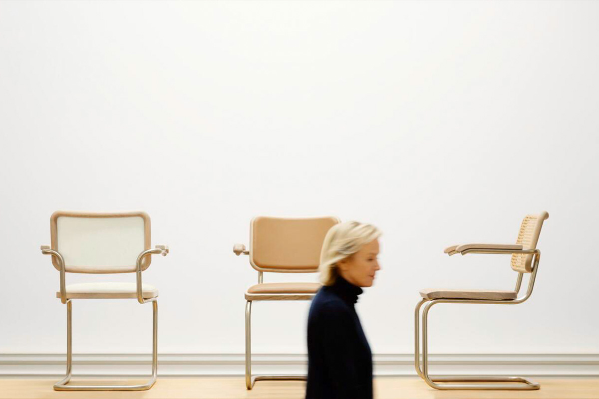 Drei Freischwinger-Stühle von JS·THONET in verschiedenen hellen Polster- und Geflechtvarianten, minimalistisch inszeniert vor weißer Wand – im Vordergrund eine vorbeigehende Frau in dunkler Kleidung, verschwommen aufgenommen. Designklassiker modern interpretiert von Jil Sander.