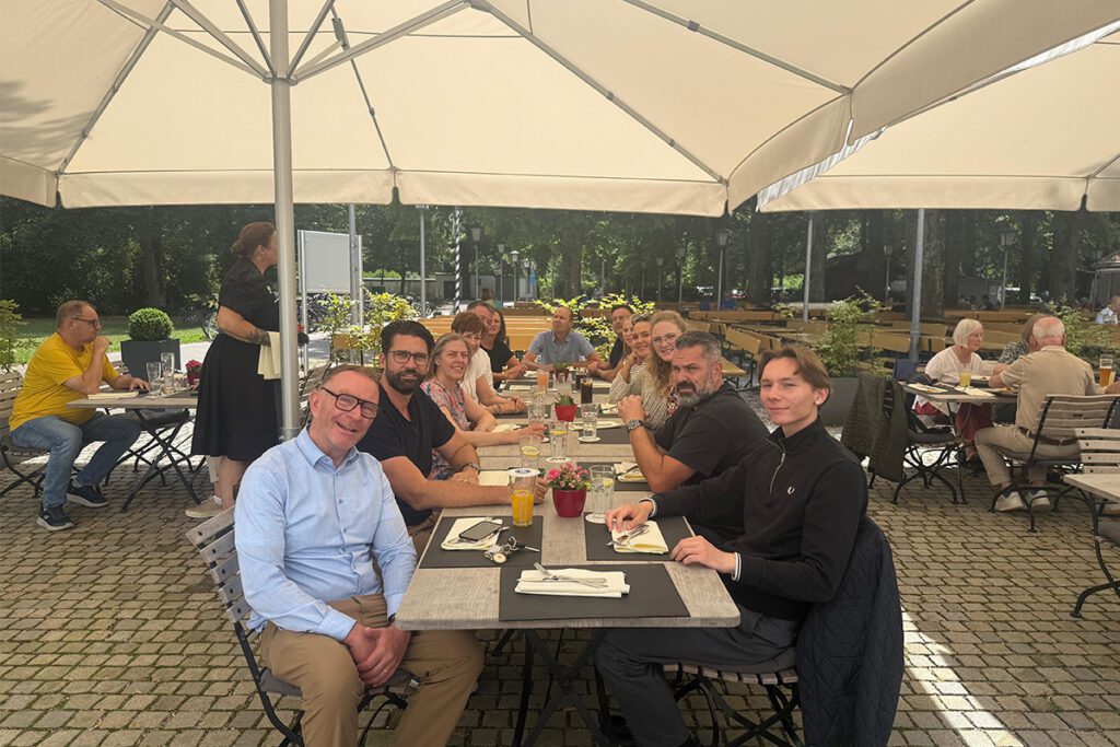 Das Hauser Team und De'berenn sitzen gemeinsam an einem langen Tisch im Außenbereich eines Restaurants unter großen Sonnenschirmen. Die Gruppe lächelt in die Kamera, das sommerliche Wetter und die entspannte Atmosphäre unterstreichen das freundschaftliche Miteinander.