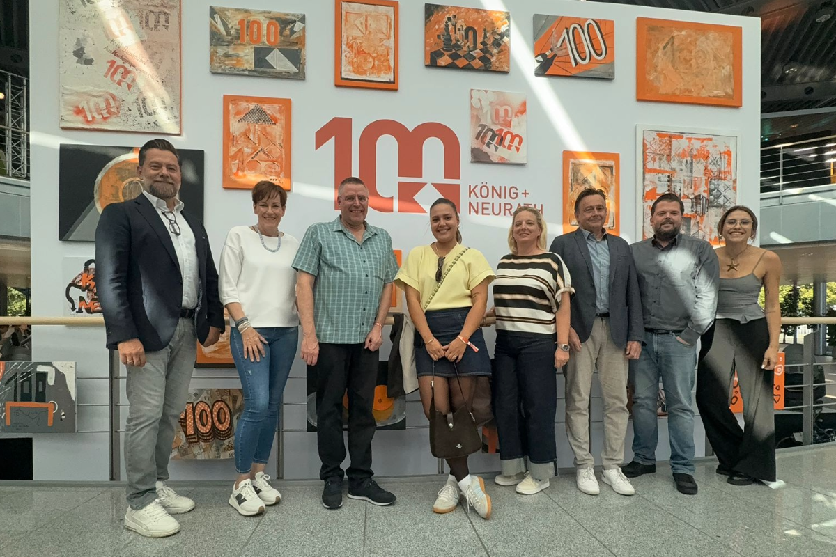 Neun Personen stehen lächelnd vor einer Wand mit gemalten Kunstwerken und dem zentralen Logo „100 Jahre König + Neurath“, das in Rot und Orange gestaltet ist.